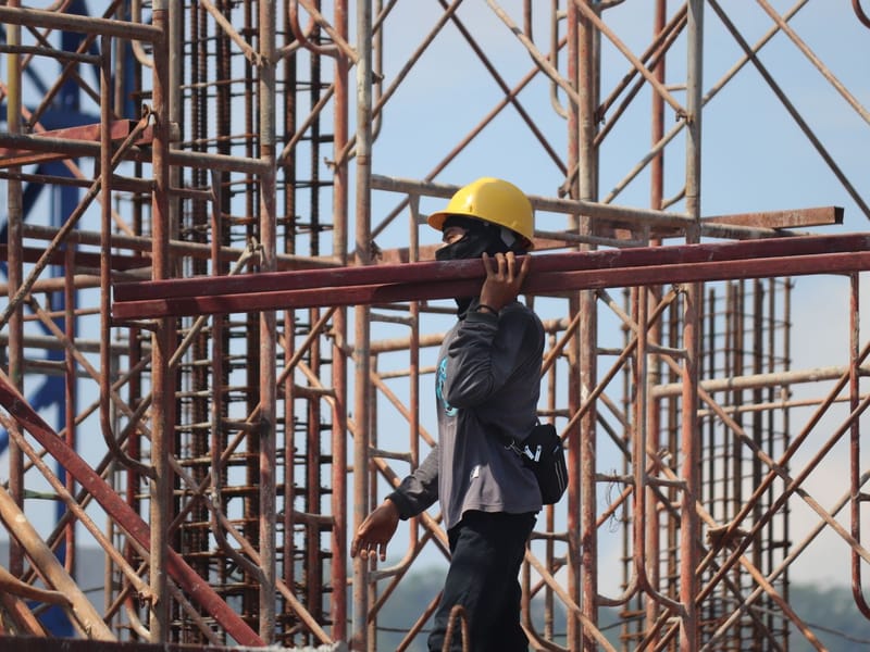 Construction labourer in a hard hat carrying steel pipes through scaffolding