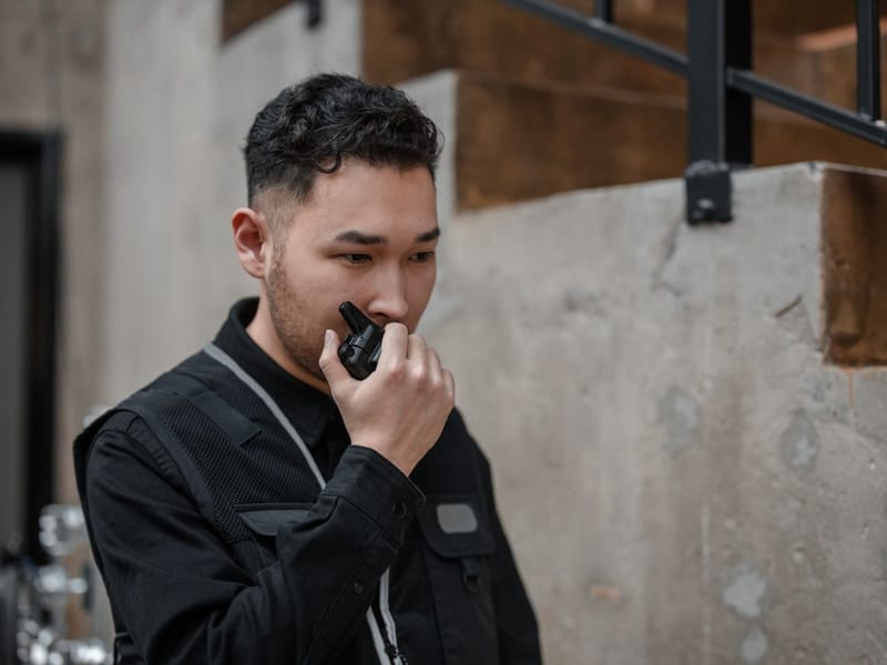 Security guard in a black uniform vest speaking into a two-way radio